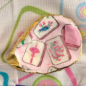 Puede incluir: Una concha de ostra decorativa con un patr&oacute;n de decoupage de fichas de mahjong sobre un fondo rosa claro con estampado geom&eacute;trico. Las fichas muestran ilustraciones de un p&aacute;jaro azul, un abanico rosa y un drag&oacute;n rosa. Los bordes est&aacute;n pintados de oro.
