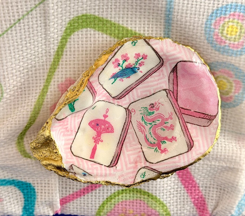 Puede incluir: Una concha de ostra decorativa con un patr&oacute;n de decoupage de fichas de mahjong sobre un fondo rosa claro con estampado geom&eacute;trico. Las fichas muestran ilustraciones de un p&aacute;jaro azul, un abanico rosa y un drag&oacute;n rosa. Los bordes est&aacute;n pintados de oro.