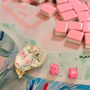 K&ouml;nnte beinhalten: Dekorative Austernschale mit goldenem Rand, bedruckt mit Mahjong-Steinen und floralen Mustern. Rosa Mahjong-Steine und W&uuml;rfel mit wei&szlig;en Punkten liegen auf einer rosa-blau gemusterten Oberfl&auml;che.