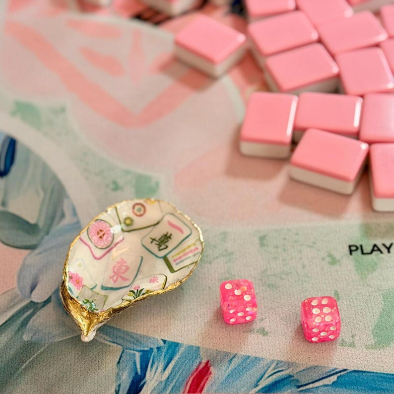 K&ouml;nnte beinhalten: Dekorative Austernschale mit goldenem Rand, bedruckt mit Mahjong-Steinen und floralen Mustern. Rosa Mahjong-Steine und W&uuml;rfel mit wei&szlig;en Punkten liegen auf einer rosa-blau gemusterten Oberfl&auml;che.