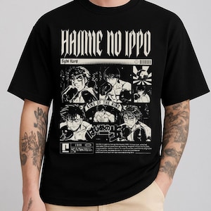 May include: Black graphic t-shirt featuring manga-style illustrations of boxers. The shirt has the text "Hajime no Ippo" and "Fight Hard" in white. The design includes multiple panels with boxing scenes and Japanese text. The shirt is worn by a person.