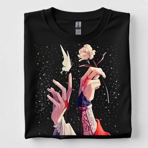 May include: Black t-shirt with an artistic illustration of hands, a butterfly, and a rose against a starry backdrop. The hands have red detailing, the butterfly is white, and the rose is white with a dark stem. A unique design for a t-shirt.