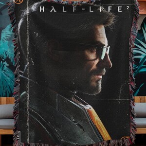Può includere: Una coperta tessuta che presenta un ritratto di profilo di un uomo con occhiali e barba, il logo di Half-Life 2 e il simbolo Lambda. Il design è prevalentemente nero con accenti arancioni e include un bordo decorativo con frange su tutti i lati.