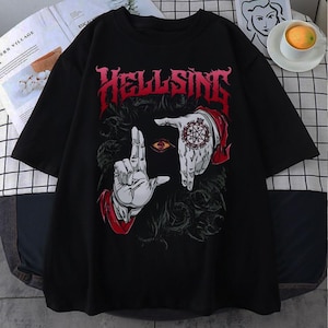 Puede incluir: Camiseta negra con la palabra "HELLSING" en letras góticas rojas. El gráfico incluye un ojo estilizado y manos con diseños intrincados, sobre un fondo oscuro. La camiseta es de manga corta, para uso casual.