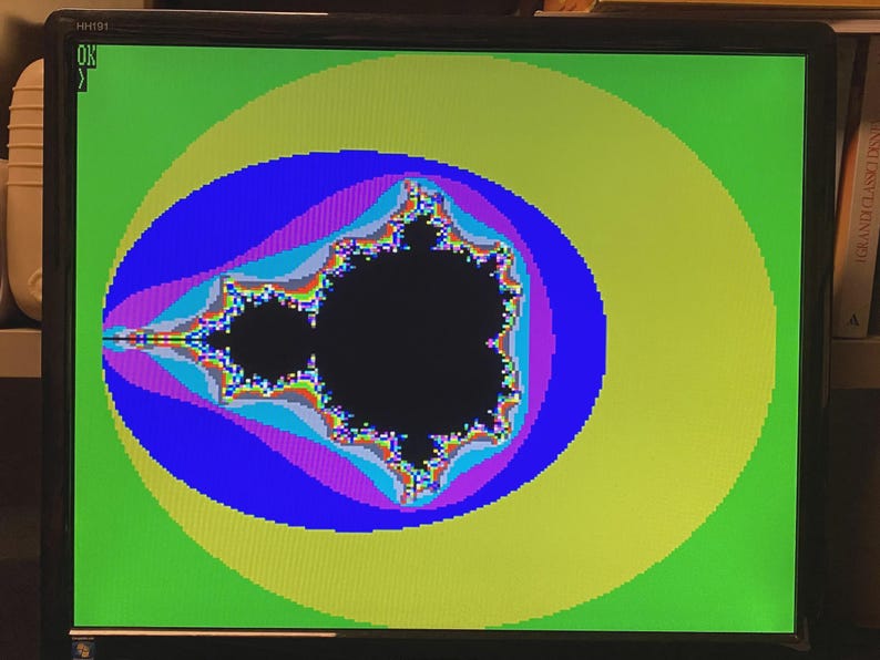 Puede incluir: Un monitor de computadora que muestra un fractal del conjunto de Mandelbrot a color. La forma negra central est&aacute; rodeada por capas conc&eacute;ntricas de azul, morado y amarillo sobre un fondo verde brillante. El texto OK es visible arriba a la izquierda.