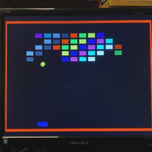 Puede incluir: Un monitor de computadora negro Hanns-G que muestra un videojuego retro de romper ladrillos. La pantalla presenta un fondo azul oscuro con una cuadr&iacute;cula de bloques rectangulares coloridos, una bola verde lima y una peque&ntilde;a paleta azul abajo.