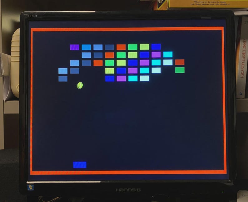 Puede incluir: Un monitor de computadora negro Hanns-G que muestra un videojuego retro de romper ladrillos. La pantalla presenta un fondo azul oscuro con una cuadr&iacute;cula de bloques rectangulares coloridos, una bola verde lima y una peque&ntilde;a paleta azul abajo.