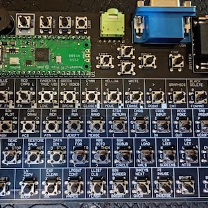 Può includere: Una tastiera a circuito stampato nero con un microcontrollore Raspberry Pi Pico verde, una porta VGA blu e un jack audio verde. La scheda è etichettata piSpectrum 2026 V 1.0 e include numerosi pulsanti tattili con etichette di testo bianche.