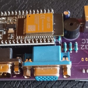 Puede incluir: Una placa de circuito impreso de color morado etiquetada como ESP32 BASIC que incluye un m&oacute;dulo microcontrolador ESP32 negro, un puerto VGA azul, un puerto serie negro, un peque&ntilde;o zumbador cil&iacute;ndrico negro, un conector dorado y componentes.