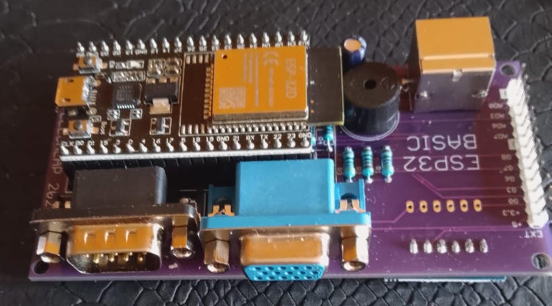 Puede incluir: Una placa de circuito impreso de color morado etiquetada como ESP32 BASIC que incluye un m&oacute;dulo microcontrolador ESP32 negro, un puerto VGA azul, un puerto serie negro, un peque&ntilde;o zumbador cil&iacute;ndrico negro, un conector dorado y componentes.