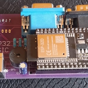 May include: A purple ESP32 BASIC printed circuit board featuring a black microcontroller module, a blue VGA port, a black serial port, a circular buzzer, a power jack, and various electronic components with white text labels printed on the board.