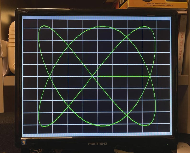 Puede incluir: Un monitor de computadora negro con el logotipo de Hanns-G en el marco inferior muestra un patr&oacute;n de curva de Lissajous verde brillante sobre un fondo de cuadr&iacute;cula blanca. La pantalla muestra una forma geom&eacute;trica compleja y sim&eacute;trica.