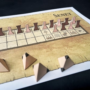 May include: Senet board game with a tan and brown color scheme. The game board features a grid with hieroglyphic symbols and the word "Senet". The game pieces are brown and pink, and the dice are tan pyramids with black accents.