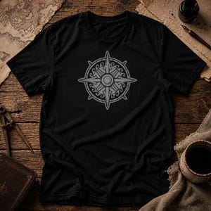May include: A black short-sleeved t-shirt featuring a centered grey graphic of a compass rose. The compass design includes four primary points, four secondary points, and a circular border decorated with a repeating wave-like pattern.