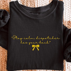 May include: Black long-sleeve shirt with the text "Stay calm, dispatcher has your back" in gold script, along with a gold bow graphic. The word "BLACK" is in the bottom right corner.