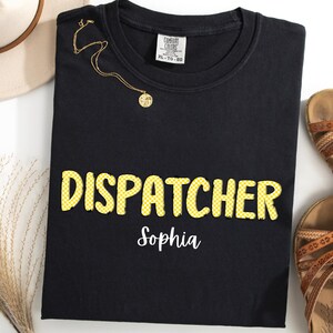 Puede incluir: Camiseta negra de cuello redondo con la palabra "DISPATCHER" en letras amarillas a cuadros y el nombre "Sophia" en blanco. Un collar dorado y un sombrero beige están al lado de la camiseta. Sandalias marrones a la derecha.