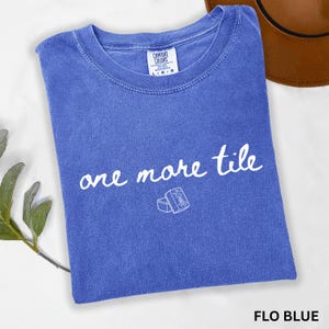 May include: A blue short-sleeved t-shirt with a crew neckline and a Comfort Colors label. The front features the white text 'one more tile' in a cursive script, positioned above a small white line drawing of a single rectangular tile.