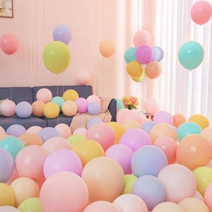 Puede incluir: Una colección de globos coloridos en varios tonos pastel, incluyendo rosa, amarillo, azul y melocotón. Algunos globos están agrupados, mientras que otros flotan libremente. La escena sugiere un ambiente festivo o de celebración.