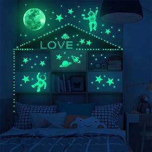 K&ouml;nnte beinhalten: Leuchtende Wandaufkleber mit Sternen, Planeten, einer Rakete, einem UFO und Astronauten. Die Aufkleber sind in Form eines Hauses angeordnet, mit dem Wort "LOVE" in der Mitte.