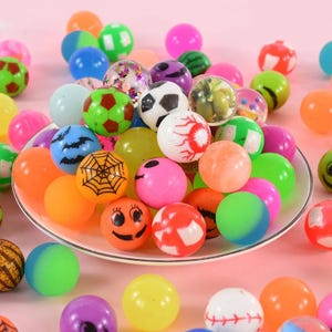 May include: A collection of colorful bouncy balls with various designs, including smiley faces, spiderwebs, and sports balls. The balls are arranged on a white plate.