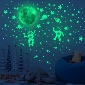 K&ouml;nnte beinhalten: Leuchtende Wandaufkleber mit einem Mond, Sternen, einer Rakete, einem Raumschiff und zwei Astronauten. Die Aufkleber befinden sich an einer blauen Wand.