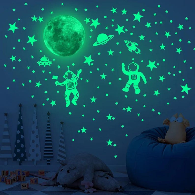 K&ouml;nnte beinhalten: Leuchtende Wandaufkleber mit einem Mond, Sternen, einer Rakete, einem Raumschiff und zwei Astronauten. Die Aufkleber befinden sich an einer blauen Wand.