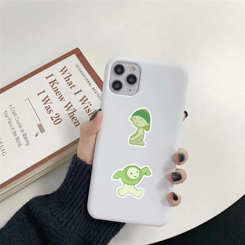 Peut inclure: Coque de t&eacute;l&eacute;phone iPhone blanche avec deux autocollants de personnages de dessin anim&eacute; verts. Les autocollants repr&eacute;sentent une cr&eacute;ature verte avec un chapeau vert et une cr&eacute;ature verte &agrave; t&ecirc;te ronde et les bras tendus.