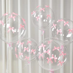 May include: Five clear, round balloons decorated with small pink bows. The balloons are inflated and appear to be made of a transparent material. The bows are evenly spaced across the surface of the balloons, creating a festive and elegant look.