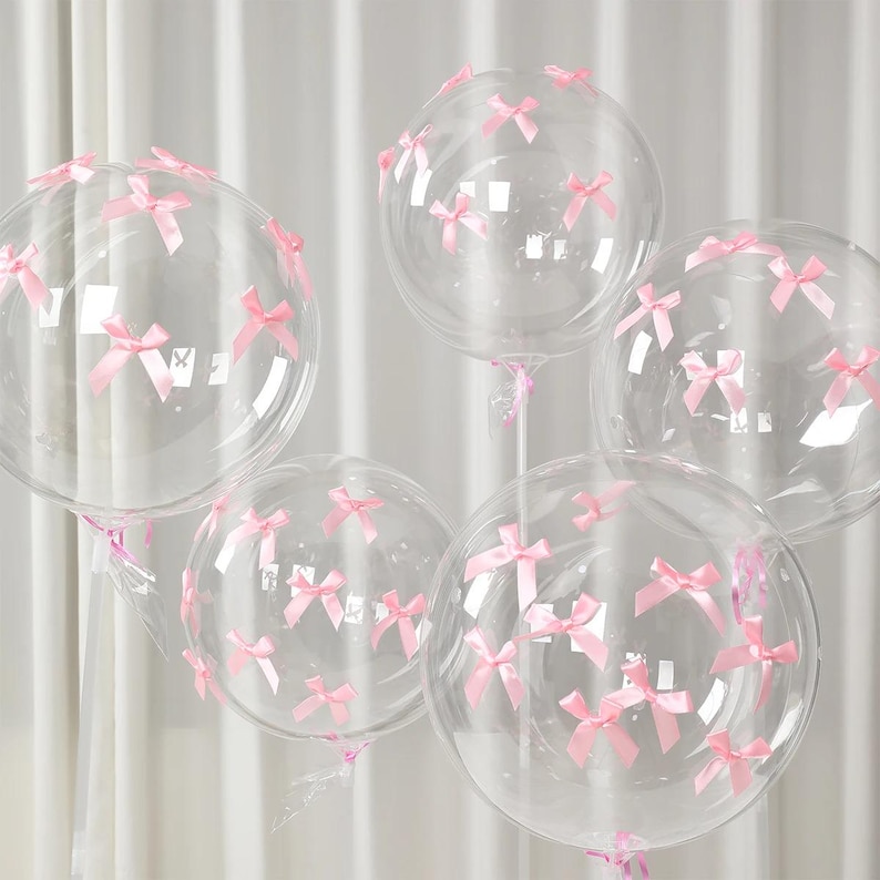 May include: Five clear, round balloons decorated with small pink bows. The balloons are inflated and appear to be made of a transparent material. The bows are evenly spaced across the surface of the balloons, creating a festive and elegant look.