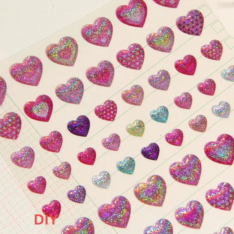May include: A sheet of iridescent heart-shaped stickers with glitter. The stickers are in various shades of pink, purple, and blue. The sheet has a grid pattern and the word "DIY" is printed in the bottom left corner.
