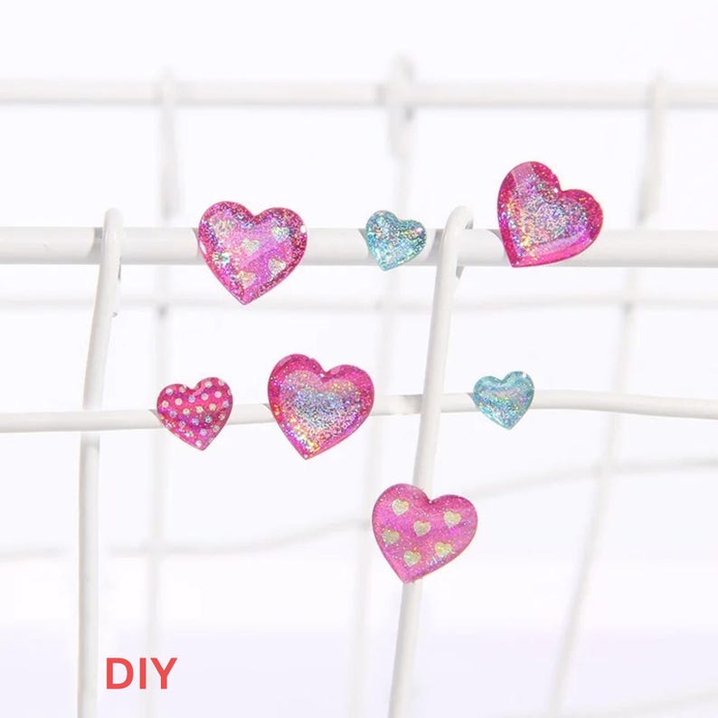 May include: Six heart-shaped earrings in a variety of colors and designs. The earrings are displayed on a white wire rack. The earrings are pink, blue, and have glitter and polka dots. The text "DIY" is visible at the bottom of the image.