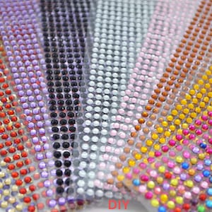 May include: A roll of adhesive-backed rhinestones in various colours, including red, orange, yellow, green, blue, purple, black, white, and silver. The rhinestones are arranged in rows on a clear plastic backing.