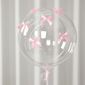 May include: A clear, round balloon decorated with small, light pink bows. The bows are evenly spaced around the balloon's surface. The balloon is attached to a clear plastic stick, suitable for party decorations.