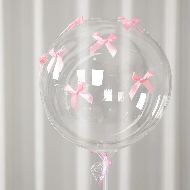 May include: A clear, round balloon decorated with small, light pink bows. The bows are evenly spaced around the balloon's surface. The balloon is attached to a clear plastic stick, suitable for party decorations.