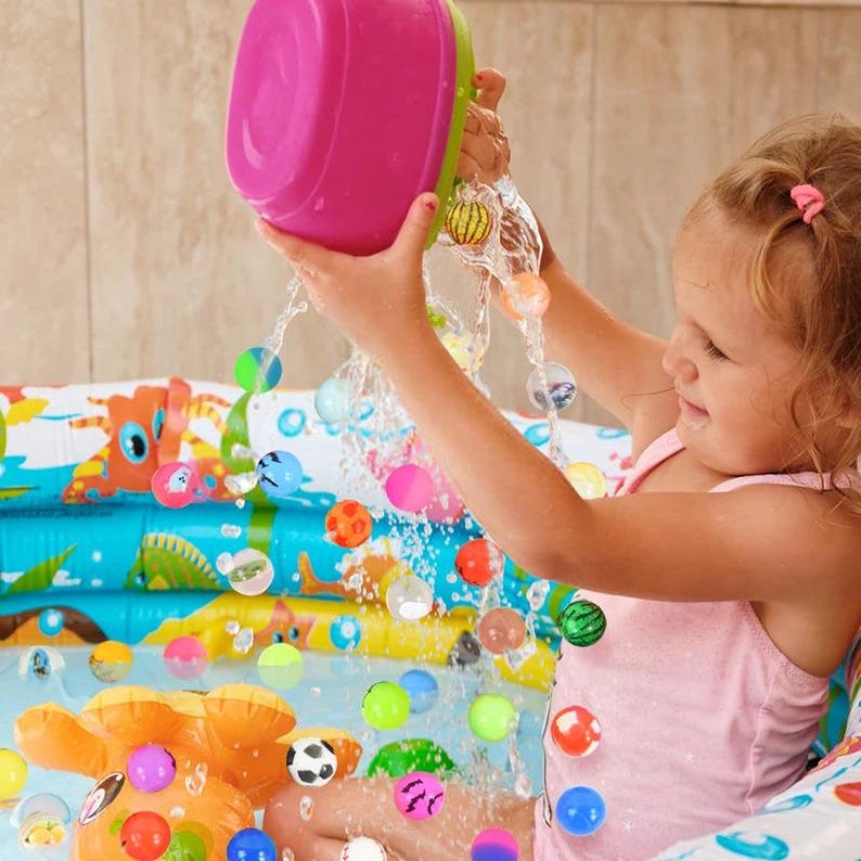 May include: A child playing in a blue and green inflatable pool with colorful bouncy balls. The child is holding a pink bowl and pouring the balls into the pool.