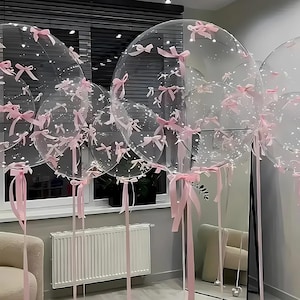 May include: Clear, round balloons decorated with small pink bows and long pink ribbons. The balloons are of varying sizes and are clustered together, creating a festive and celebratory atmosphere. Some balloons have small white dots inside.