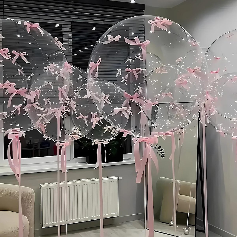 May include: Clear, round balloons decorated with small pink bows and long pink ribbons. The balloons are of varying sizes and are clustered together, creating a festive and celebratory atmosphere. Some balloons have small white dots inside.