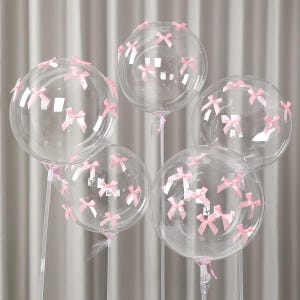 May include: Five clear, round balloons decorated with small pink bows. The balloons are attached to clear sticks and are arranged against a soft, neutral background. The bows are evenly spaced across the surface of each balloon.