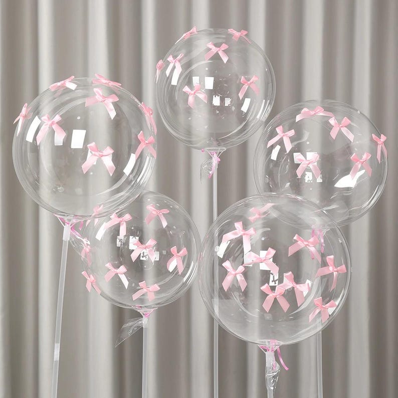 May include: Five clear, round balloons decorated with small pink bows. The balloons are attached to clear sticks and are arranged against a soft, neutral background. The bows are evenly spaced across the surface of each balloon.