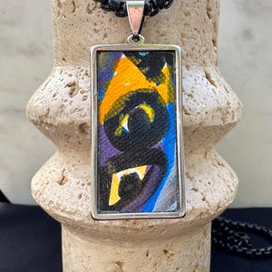 May include: A silver chain necklace with a rectangular pendant featuring abstract art in shades of yellow, blue, purple, and black.