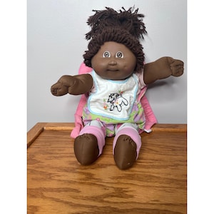 May include: A vintage Cabbage Patch Kid doll with dark skin, brown yarn hair, and brown eyes. The doll wears a white bib with a floral and dog design, a pink cape, and pink and white striped pants and shoes. The doll is sitting on a wooden surface.