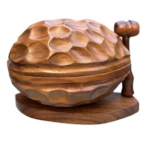 May include: A brown wooden nutcracker shaped like a walnut with a textured, dimpled surface. The object consists of a two-part walnut-shaped container resting on a flat wooden base, accompanied by a small wooden mallet attached to the side.