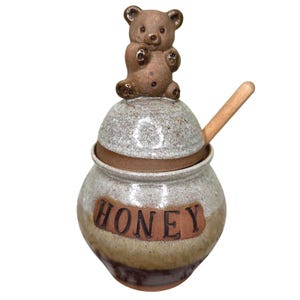 May include: A ceramic honey pot with a speckled cream and brown glaze, featuring a sculpted brown bear on the lid and the word HONEY embossed on the front. A light-colored wooden honey dipper is inserted into the side of the lid opening.