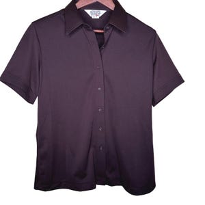 May include: A solid dark purple short-sleeve button-down shirt with a classic collar. The garment features a front button placket with matching dark purple buttons and a straight hemline. A white interior neck label displays text including 100% polyester.