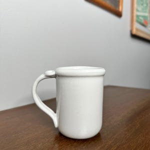 May include: A small, white ceramic mug with a rounded body and a curved handle. The mug has a simple, minimalist design, perfect for coffee or tea. The handle features a small, decorative detail where it attaches to the mug.