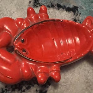 Może przedstawiać: Jasnoczerwony talerz w kształcie kraba z błyszczącym wykończeniem. Talerz ma szczegółowy wzór kraba, w tym szczypce, oczy i segmentowe ciało. Talerz jest prawdopodobnie ceramiczny lub z podobnego materiału.