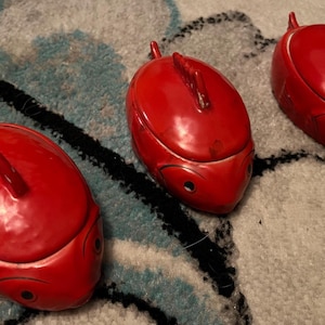 May include: Three red, oval-shaped decorative boxes with a glossy finish. Each box has a small, curved antenna-like feature on top and two small, round eye-like details on the front. The boxes are arranged on a patterned surface.