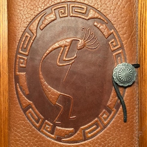 May include: A brown leather journal with a Kokopelli design embossed on the cover. The design is surrounded by a geometric border. A silver-colored button with a black cord closure is on the right side.
