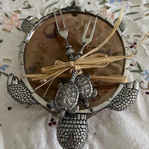 May include: A silver turtle-shaped serving dish with a brown shell-like interior. The dish features two silver forks tied together with a straw bow. The dish is decorated with three turtle figures.