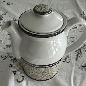 Puede incluir: Una tetera de cerámica blanca con un diseño moteado marrón y verde. La tetera tiene una tapa con un borde marrón y un pomo plateado.